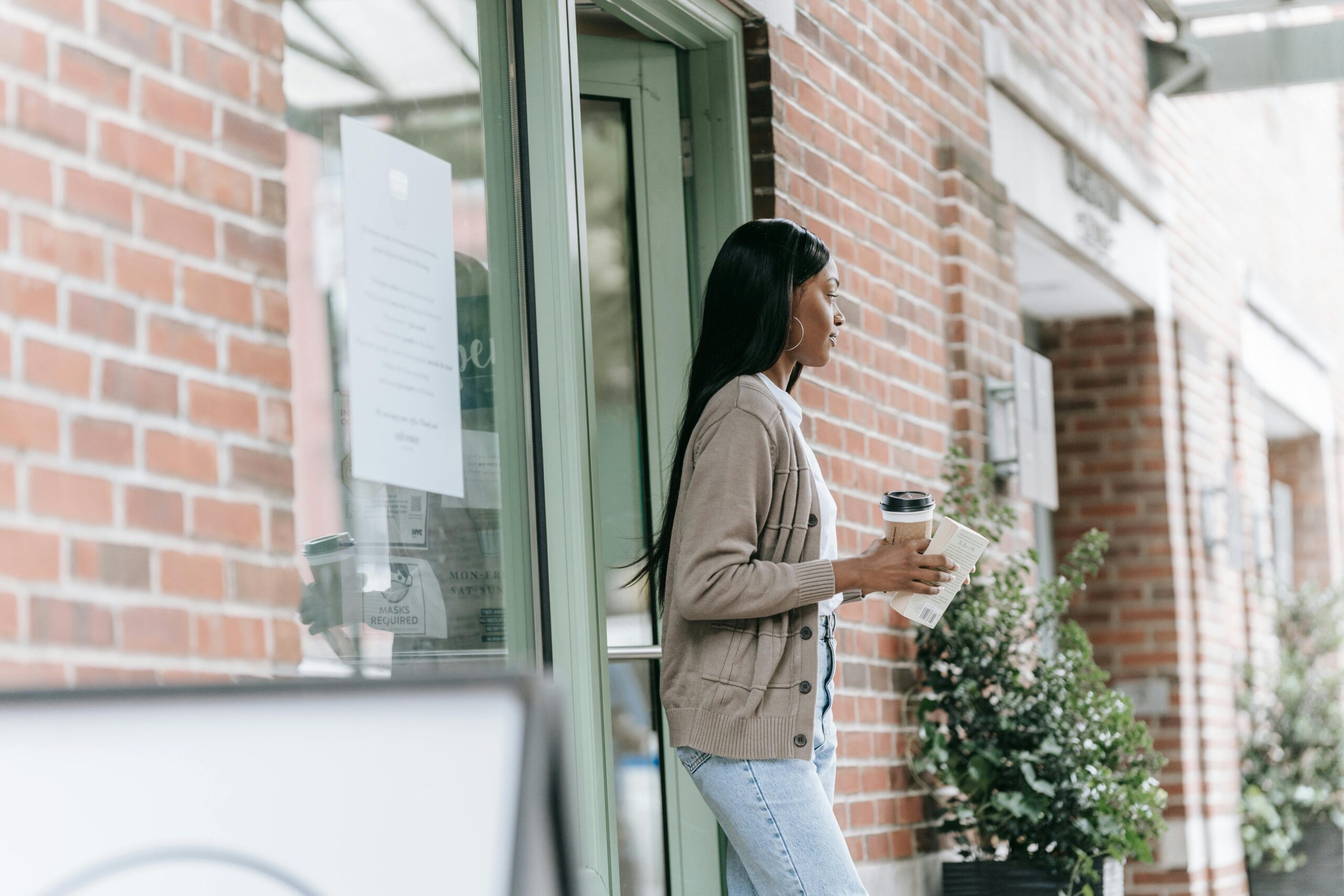 African American woman leaving a café with a coffee and book, surrounded by plants.